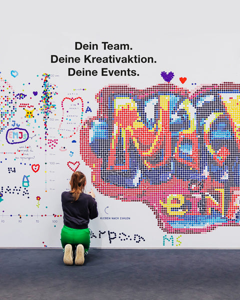 Person klebt bunte Klebepunkte auf dot on Eventwand mit Pixelartmotiv und Feedbackelementen